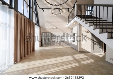 Bright, modern interior featuring a high-ceilinged room with large windows, tile flooring, and a minimalist kitchen with gray cabinets. A staircase leads to an upper level