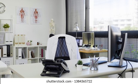 bright and modern medical doctor office complete with personal computer and telephone on desk white lab coat on chair. empty nobody in room in clinic. hospital workplace with sunshine through window.