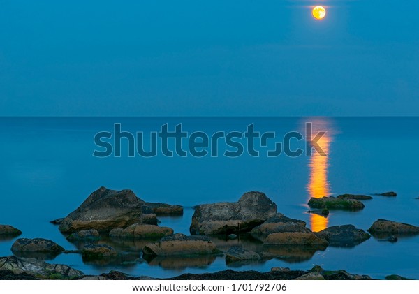Bright moon over the sea in the night sky. Lunar
path on the surface of the sea. 
Rock formations protrude from the
water.