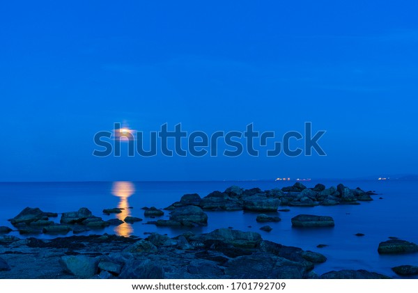 Bright moon over the sea in the night sky. Lunar\
path on the surface of the sea. \
Rock formations protrude from the\
water.