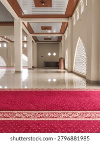 Bright mosque interior featuring red prayer carpet, elegant columns, reflective floor, and Islamic geometric windows illuminated by natural sunlight.