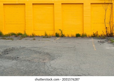 A Bright Orange Exterior Brick Wall With Garage Openings Or Bays That Have Been Enclosed With Bricks. The Foreground Is Paved With Some Weeds And Plants Growing Up Along The Edge Of The Building.