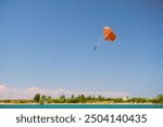 A bright orange parasail soars high in a clear blue sky over a picturesque coastal landscape. Two people dangle beneath the parasail, enjoying a thrilling adventure above the turquoise waters below.