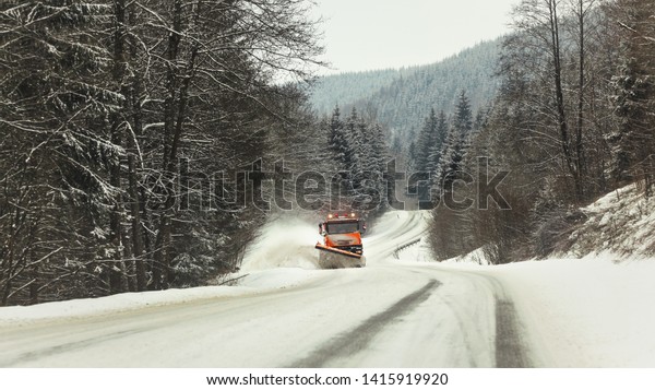 Bright orange plough truck cleaning snow on forest\
road in winter, view from car driving other way, trees around,\
overcast day