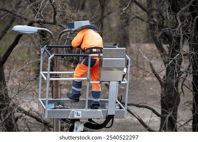 In a bright orange safety suit, worker on an elevated platform repairs streetlight surrounded by bare trees, maintenance task, outdoor scene concept - Powered by Shutterstock