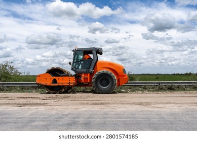 Bright orange single-drum soil compactor roller operating on a road construction site under a cloudy sky.
