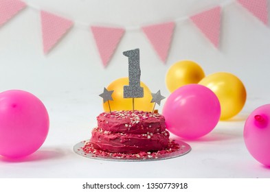 Bright pink first birthday cake with number 1 (one) on top with two stars and sprinkles, pink and gold balloon and bunting with a white background