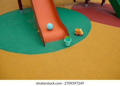 Bright playground setup featuring an orange slide, a blue dotted ball, a green bucket, and a small yellow toy truck on a multicolor rubber surface.