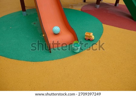 Bright playground setup featuring an orange slide, a blue dotted ball, a green bucket, and a small yellow toy truck on a multicolor rubber surface.