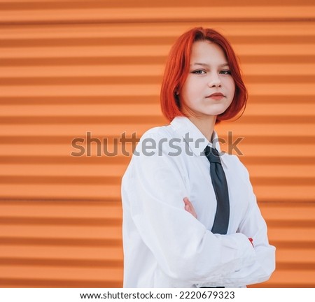 Bright portrait of teenage girl with painted red dyed hair in white school shirt and necktie standing near orange wall background with hands crossed body posture.