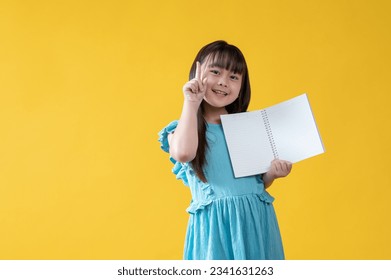 A bright and pretty little Asian girl in a blue dress is holding an opened book with empty pages, pointing her finger up, smiling, and looking at the camera. isolated yellow background - Powered by Shutterstock