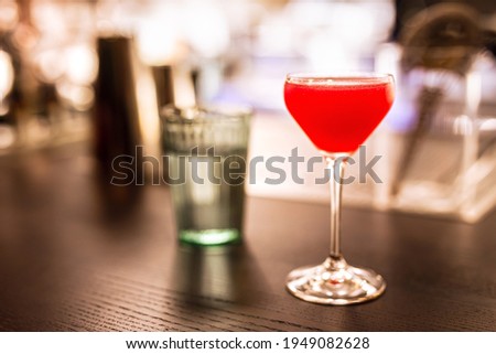 A bright red alcoholic cocktail in a coupe glass and a glass of water at the bar counter. A horizontal lifestyle photo with shallow depth of field and copy space.