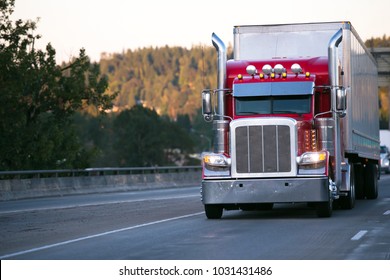 Bright red classic big rig semi truck with high exhaust pipes and chrome accessories transporting goods in dry van trailer move on evening road with turn on headlight