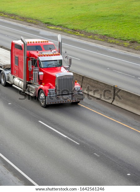 Bright Red Classic Big Rig diesel Semi Truck with\
windows on the cab roof and Chrome Accessories and Exhaust Pipes\
Running with Empty Flat Bed Semi Trailer on the Multilines Divided\
Highway Road