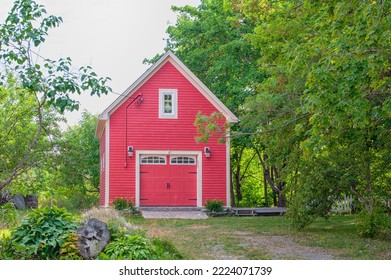 A Bright Red Country Style Red Barn Or Single Car Garage Surrounded By Lush Green Trees And Shrubs. The Historic Building Has A Small Window On The Top Floor And A Garage Door On The Lower Level.