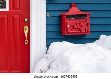 A Bright Red Decorative Metal Mailbox Or Letterbox On A Blue Exterior Wall Of A House With A Red Door, Brass Handle And White Trim. The Mailbox Has A Mound Of Fresh Snow In Front Of The Building. 