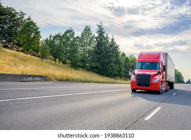 Bright red diesel big rig industrial professional semi truck with black grille transporting commercial cargo in dry van semi trailer driving on the straight wide highway road with trees on hillside