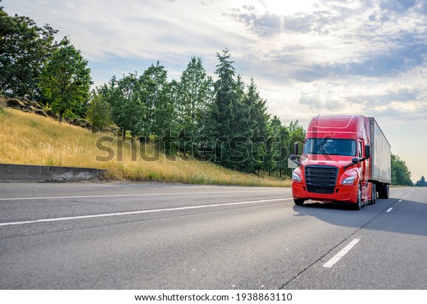 Bright red diesel big rig industrial\
professional semi truck with black grille transporting commercial\
cargo in dry van semi trailer driving on the straight wide highway\
road with trees on\
hillside