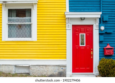 A bright red door of a building with blue and yellow wooden clapboard walls. The house has a red decorative metal mailbox. There's a green shrub near the red door with white trim.  - Powered by Shutterstock