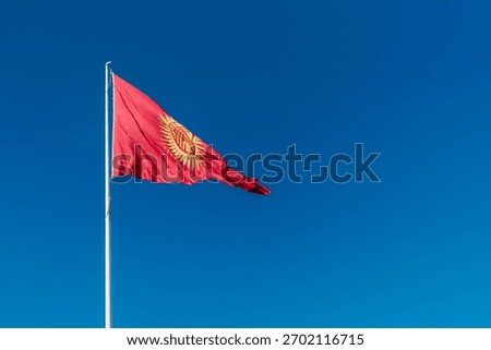 Bright red flag featuring a radiant golden sun with forty rays and a central tunduk emblem, fluttering on a high flagpole, symbolizes unity, tradition, and independence of Kyrgyzstan