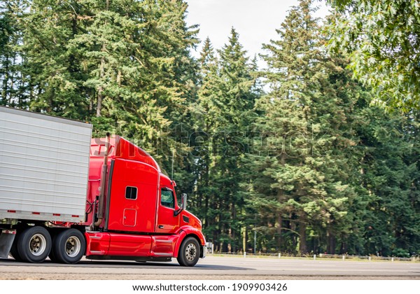 Bright red long hauler big rig red industrial semi\
truck transporting frozen and chilled foods in refrigerator semi\
trailer running on the straight highway road with green trees on\
the side