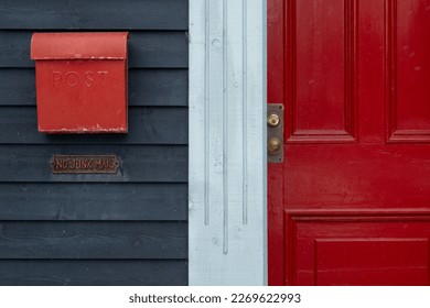 A bright red metal mailbox or letterbox on a deep blue wooden wall with a red door.  The mailbox has a curved shape cover. There's a shiny door handle, brass metal. The trim house is white in color.