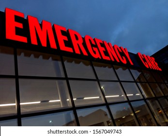 Bright Red Neon Emergency Care Sign Outside A Hospital Building At Night Against A Dark Blue Sky Above Shiny Reflections On Glass Plate Windows