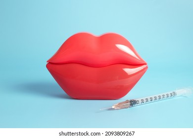 Bright red plastic female lips and a filler and hyaluronic acid injection syringe on a light blue background.
