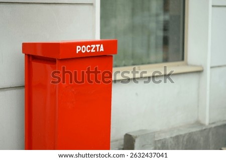 A bright red post box labeled Poczta is positioned next to a white building, with natural light illuminating the surroundings.