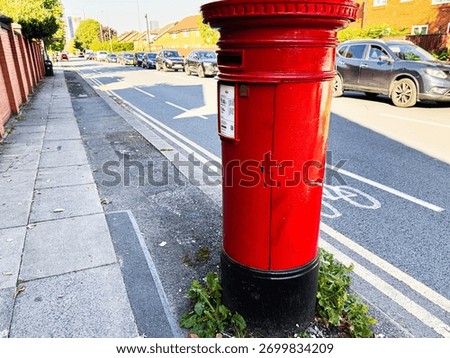 Bright red post box on a Manchester sidewalk, with parked cars and graffiti nearby, creating urban street scene.