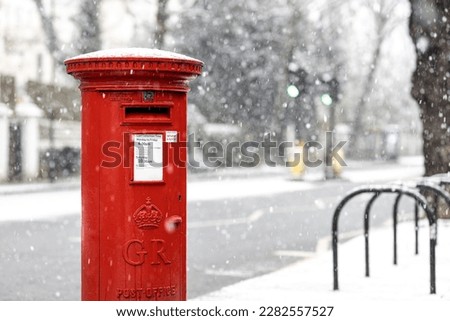Bright red post box stands out on a freshly snow covered London street beside townhouses; ideal for winter travel campaigns, Christmas mailers, features and British city branding
