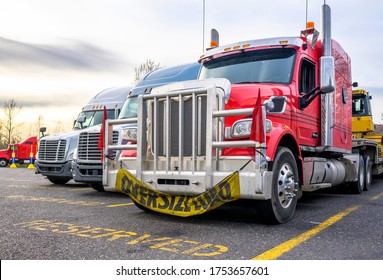 Bright Red Powerful Big Rig Semi Truck With Oversize Load Sign On The Grill Guard Bumper Standing With Loaded Semi Trailer At Row On The Truck Stop With Another Semi Trucks On Reserved Parking Spots