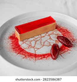 A bright red rectangular fruit terrine plated on a white ceramic dish, set on a fine web of red sauce with dark garnish alongside. Soft natural light picks out the clean edges and translucent surface of the dessert. A restaurant food image for dessert menus, patisserie and fine dining themes.