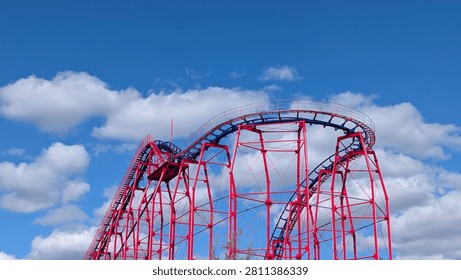 A bright red roller coaster arcs against a clear blue sky with fluffy clouds.