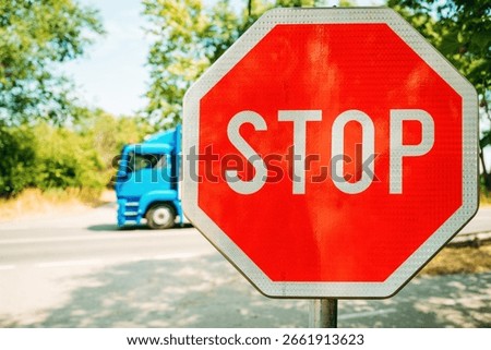 Bright red stop sign in sharp focus on a sunny day, with a blurred blue truck passing by in the background. Set against green trees and a quiet road. Selective focus.