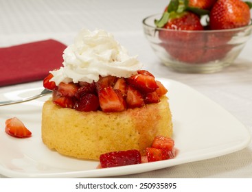 Bright Red Strawberries Topped With Syrup And Whipped Cream On A Yellow Shortcake. Served On A Square Plate Set On A White Table Cloth. Bowl Of Strawberries In The Background.