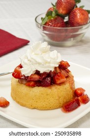 Bright Red Strawberries Topped With Syrup And Whipped Cream On A Yellow Shortcake. Served On A Square Plate Set On A White Table Cloth. Bowl Of Strawberries In The Background.