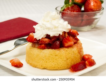 Bright Red Strawberries Topped With Syrup And Whipped Cream On A Yellow Shortcake. Served On A Square Plate Set On A White Table Cloth. Bowl Of Strawberries In The Background.