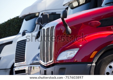 Bright red and White Big rigs semi trucks of different makes and models stand in row on truck stop parking lot for truck driver rest according to logbook delivery schedule