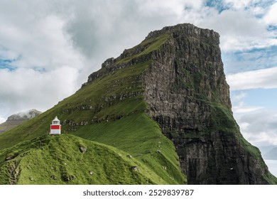 A bright red and white lighthouse sits on a lush green hill, framed by a towering rocky mountain under a partly cloudy sky. The scene is serene and picturesque. - Powered by Shutterstock