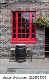 Bright red window in a red brick wall of a typical old London pub with a hanging basket of flowers and a beer barrel and stools - Shutterstock ID 2208583305