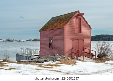 A bright red wooden building near a beach overlooking the ice blue ocean and thick cloudy sky. The storage building has a small glass window and door painted white. There's white snow on the ground.  - Powered by Shutterstock