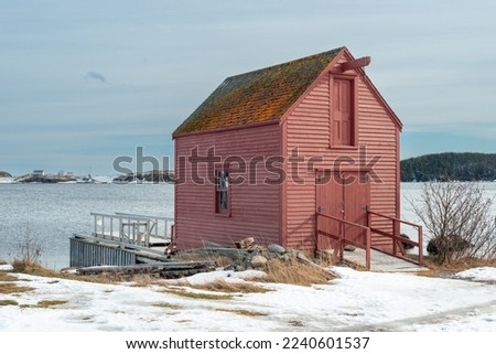 A bright red wooden building near a beach overlooking the ice blue ocean and thick cloudy sky. The storage building has a small glass window and door painted white. There's white snow on the ground. 