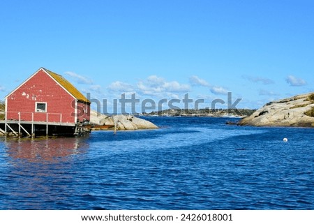 A bright red wooden storage shed with a peaked roof and small glass window on the shoreline of Peggy's Cove, Nova Scotia. The vintage fishing building is built on stilts over the deep blue water.