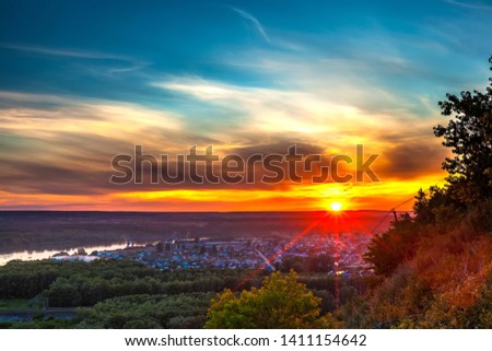 Bright red yellow orange setting sun on a blue sky with dramatic dark clouds over the city on the banks of the Belaya River in a green dense forest. Sunset over Ufa, Bashkortostan, Russia.