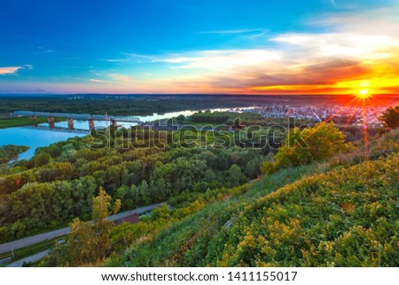 Bright red yellow orange setting sun on a blue sky from a hillside, the valley of the Belaya river in a green dense forest and a metal railway bridge. Sunset over Ufa, Bashkortostan, Russia.