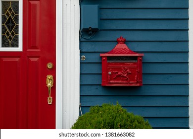 A bright retro red metal mailbox, or letterbox, is affixed to the exterior wall of a blue wooden house made of clapboard. There's a red door with a gold door knob and white trim on the building.