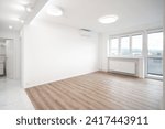 A Bright, spacious room with white walls, two ceiling lights, wooden flooring, and large windows. An open doorway leads to a tiled kitchen and bathroom area.”