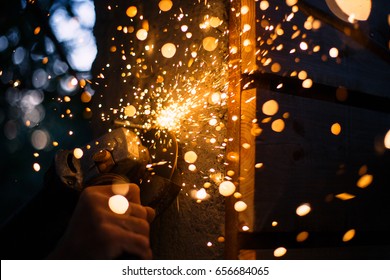 Bright sparks are flying from cutting metal abrasive disk of the angle grinder. Man's hands with a grinder are cutting the metal rod. A man is building a fence from wood and concrete at outdoor.