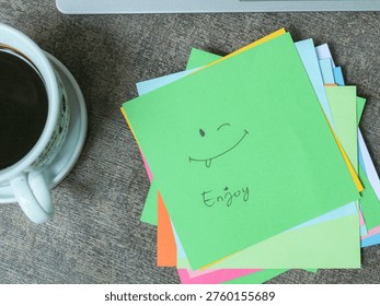 Bright sticky notes featuring a smiling face and enjoy message on a wooden desk with coffee cup and laptop creating a cheerful workspace atmosphere perfect for creative and motivational content.